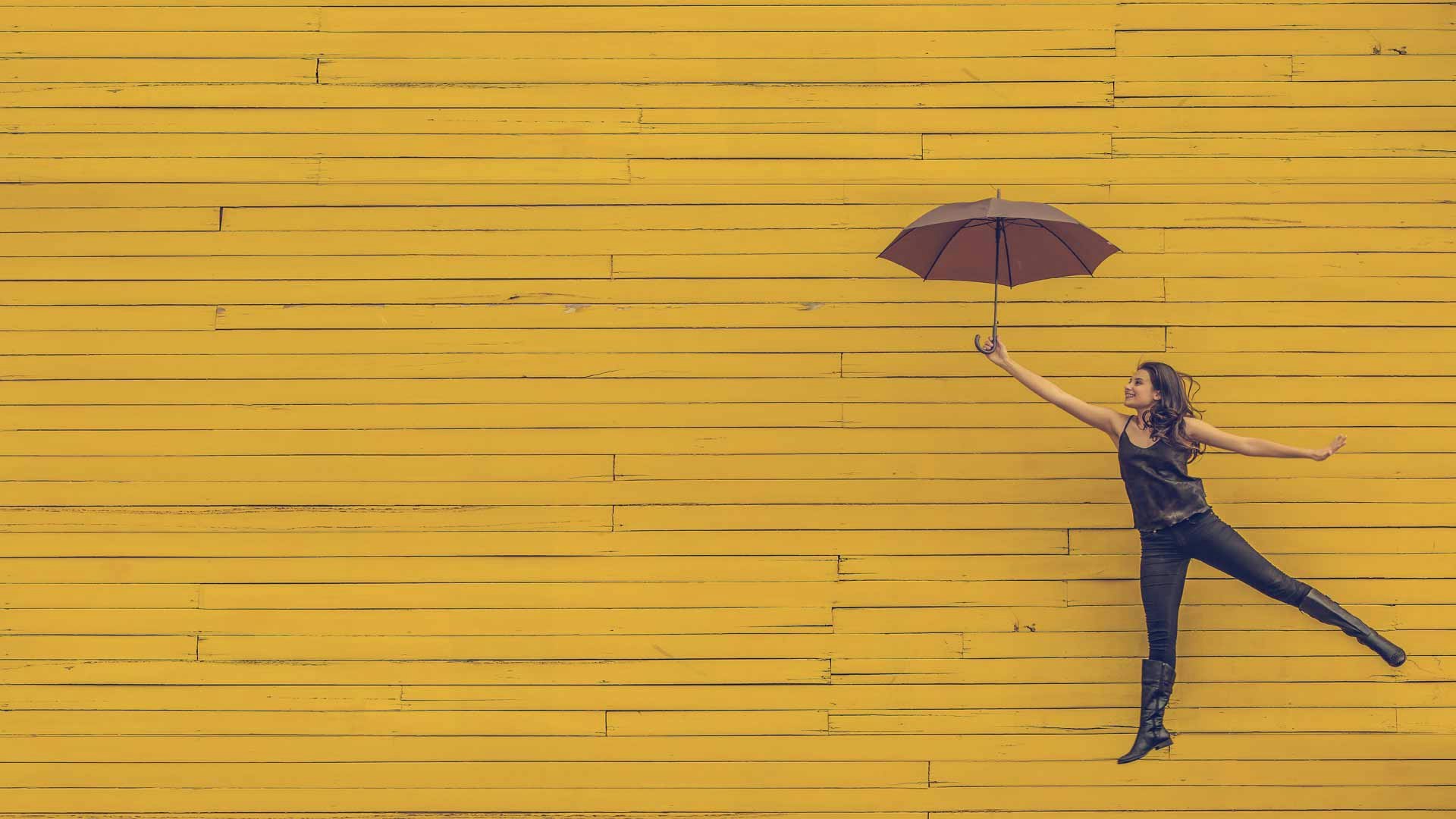 Young woman floating away with an umbrella against a yellow wooden wall